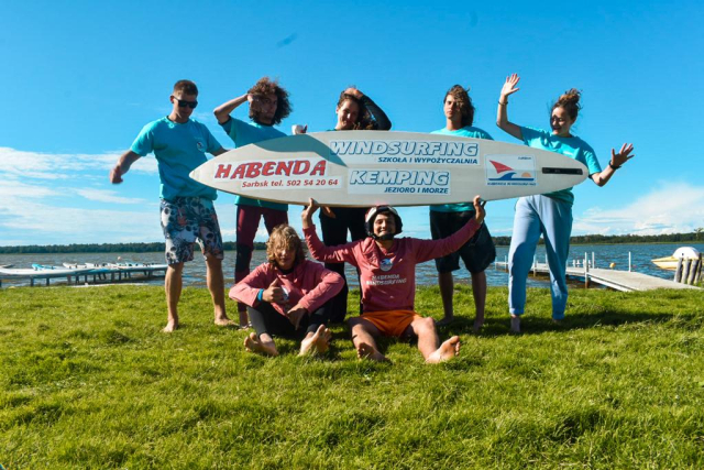 Grupa młodych osób z deską windsurfingową i banerem szkoły na trawie.